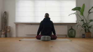 Ein Bild einer Frau, die in einem Yoga-Studio auf einer Yogamatte sitzt und in einer meditativen Haltung die Augen geschlossen hat. Ihre Hände ruhen auf den Knien, während sie gelassen und konzentriert wirkt, die Praxis der Achtsamkeit und inneren Ruhe verkörpernd.